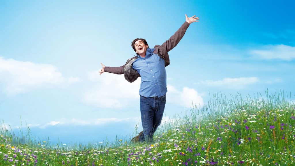 Jim Carrey, les bras écartés et le sourire aux lèvres, court dans un champ fleuri sous un ciel bleu dans le film Yes Man.