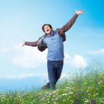 Jim Carrey, les bras écartés et le sourire aux lèvres, court dans un champ fleuri sous un ciel bleu dans le film Yes Man.