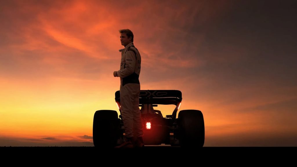 Silhouette du pilote Sonny Hayes (Brad Pitt) debout devant sa monoplace Apex GP sur un circuit, avec un ciel de coucher de soleil orange intense en arrière-plan.