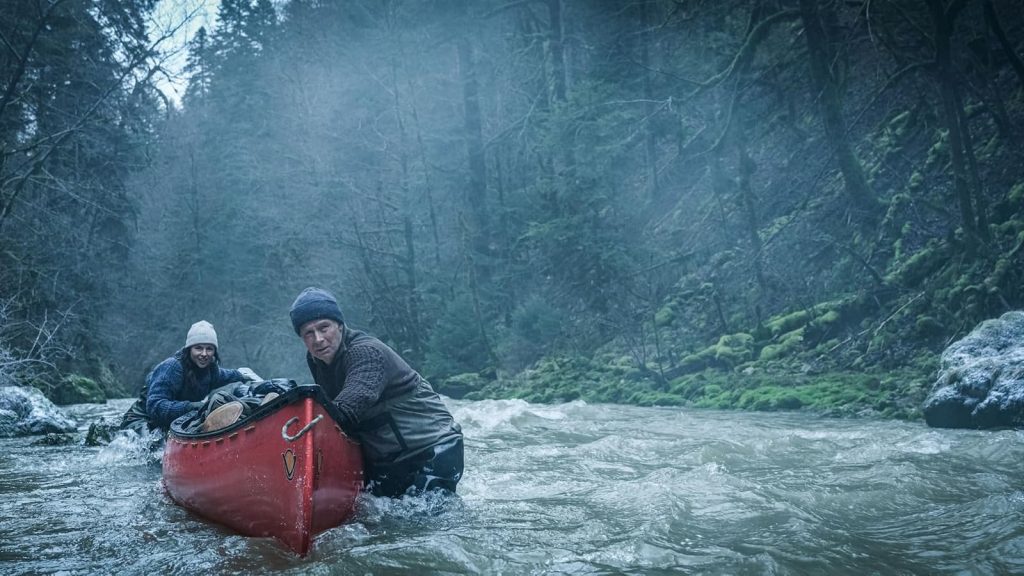 Franck Dubosc, portant un bonnet bleu, et Laure Calamy, portant un bonnet blanc, sont debout dans l'eau glaciale d'un torrent de montagne, le visage tendu et inquiet. Ils encadrent un canoë rouge contenant un cadavre. En arrière-plan, une forêt de conifères dense et couverte de mousse verte s'élève, sous un ciel brumeux et froid. La scène dégage une atmosphère de tension et de survie.