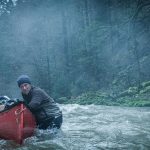 Franck Dubosc, portant un bonnet bleu, et Laure Calamy, portant un bonnet blanc, sont debout dans l'eau glaciale d'un torrent de montagne, le visage tendu et inquiet. Ils encadrent un canoë rouge contenant un cadavre. En arrière-plan, une forêt de conifères dense et couverte de mousse verte s'élève, sous un ciel brumeux et froid. La scène dégage une atmosphère de tension et de survie.