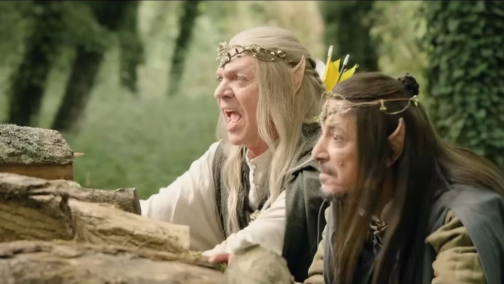 Plan moyen de deux hommes costumés en elfes de fantaisie, accroupis derrière une pile de bûches dans une forêt. L'homme de gauche (Marc Labrèche), avec de longs cheveux blonds, crie énergiquement la bouche grande ouverte. L'homme de droite (Hafid F. Benamar), aux cheveux bruns, regarde attentivement devant lui avec une expression sérieuse. Tous deux portent des oreilles pointues prosthétiques et des diadèmes détaillés.