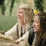 Plan moyen de deux hommes costumés en elfes de fantaisie, accroupis derrière une pile de bûches dans une forêt. L'homme de gauche (Marc Labrèche), avec de longs cheveux blonds, crie énergiquement la bouche grande ouverte. L'homme de droite (Hafid F. Benamar), aux cheveux bruns, regarde attentivement devant lui avec une expression sérieuse. Tous deux portent des oreilles pointues prosthétiques et des diadèmes détaillés.
